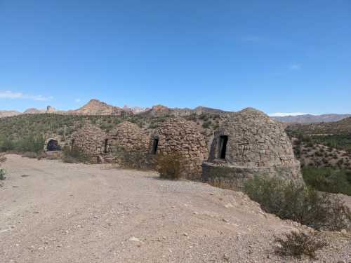 Stone kilns stand in a desert landscape, surrounded by sparse vegetation and mountains under a clear blue sky.