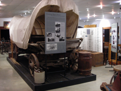 A covered wagon displayed in a museum, with informational panels and artifacts in the background.