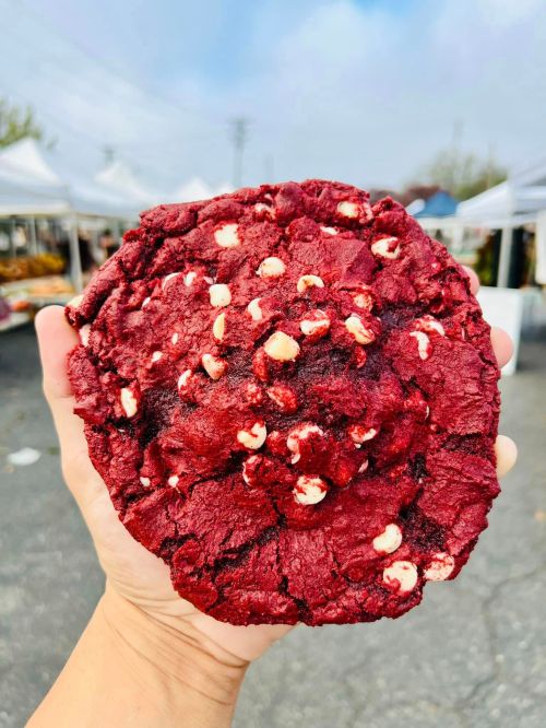 A hand holding a large red velvet cookie with white chocolate chips, set against a market backdrop.