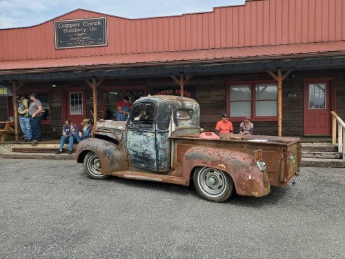 A vintage, rusty pickup truck parked in front of a rustic building with people sitting nearby.