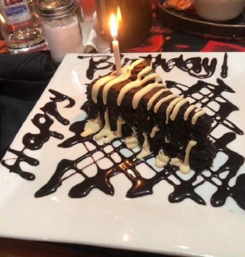 A slice of birthday cake with a candle, drizzled with chocolate and cream, on a plate that says "Happy Birthday."