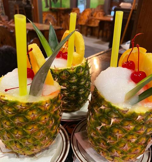 Three tropical drinks served in hollowed-out pineapples, topped with cherries and pineapple slices, with colorful straws.