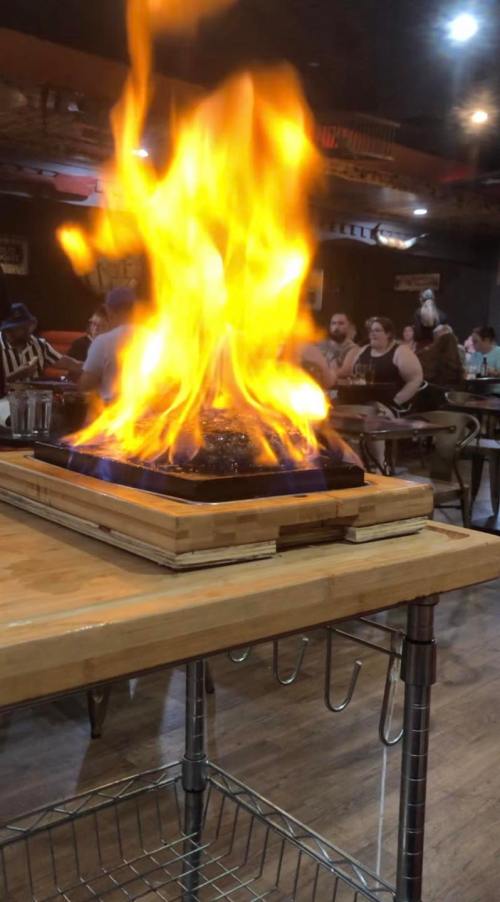 A vibrant flame rises from a grill on a wooden table, with diners enjoying their meals in the background.