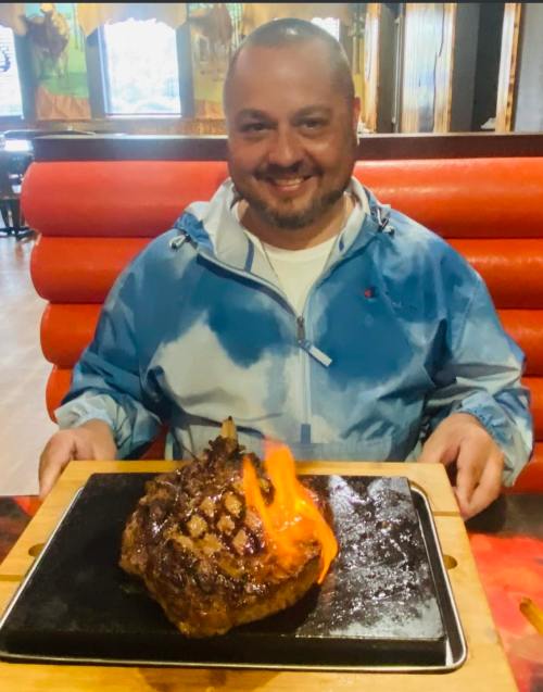 A smiling man holds a large steak on a sizzling platter with flames, in a restaurant setting.
