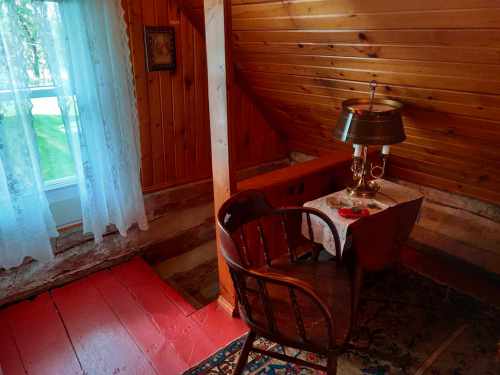 Cozy wooden room with a chair, small table, lamp, and a window with lace curtains. Warm, rustic atmosphere.
