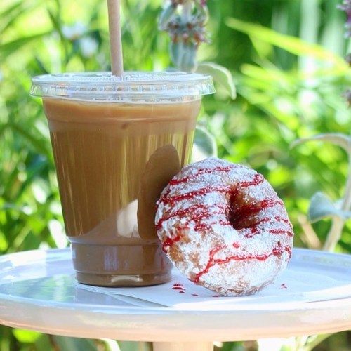 A clear cup of iced coffee next to a powdered donut drizzled with red icing, set on a white table outdoors.