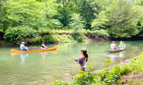 Two canoes on a river with people fishing and enjoying a sunny day surrounded by lush greenery.