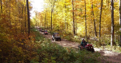 A line of ATVs travels down a dirt path through a vibrant autumn forest with colorful leaves.