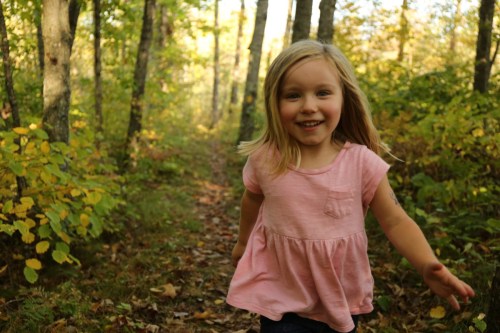 A young girl in a pink shirt runs joyfully along a forest path surrounded by trees and autumn foliage.