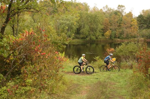 Two cyclists ride along a grassy path near a calm lake, surrounded by trees with autumn foliage.