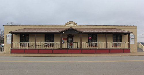 A single-story building with a tan exterior, red accents, and a covered porch, featuring a "Welcome" sign and neon lights.