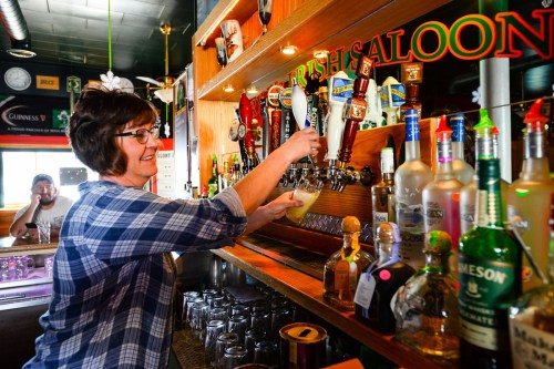 A bartender pours a drink from a tap at an Irish saloon, with various bottles displayed behind her.