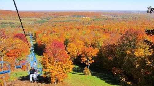 A scenic view from a ski lift overlooking vibrant autumn foliage in various shades of orange, red, and yellow.