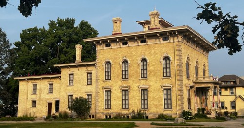 Historic yellow mansion with ornate architecture, large windows, and a lush green backdrop.