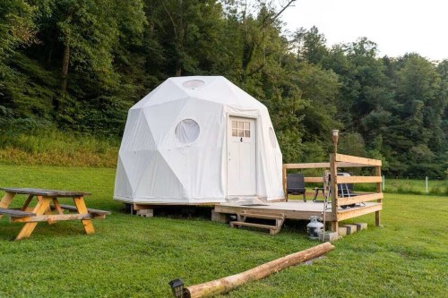 A white geodesic dome on a wooden deck, surrounded by green grass and trees, with a picnic table nearby.