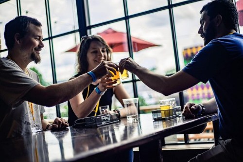 Three friends clink glasses over a table, enjoying drinks in a lively indoor setting with large windows.