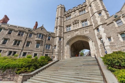Stone archway of a historic university building, flanked by greenery and steps leading up to the entrance.