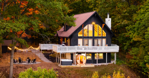 Modern dark cabin with a lit-up deck, hot tub, and barrel sauna nestled in fall trees in Michigan.