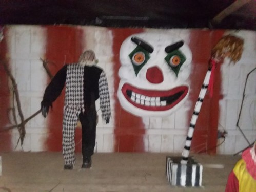 A creepy clown figure stands next to a large, menacing clown face on a red and white wall.