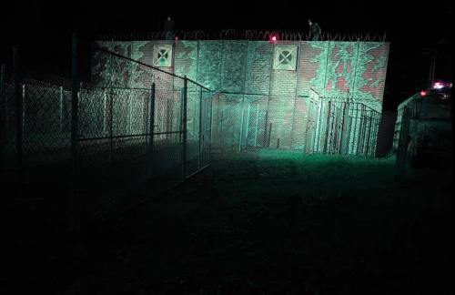 A dimly lit, fenced area at night with a brick wall and barbed wire, illuminated by green light.