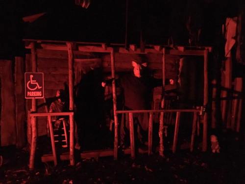 A dimly lit scene of a rustic wooden structure with a figure inside, surrounded by eerie shadows and a parking sign.