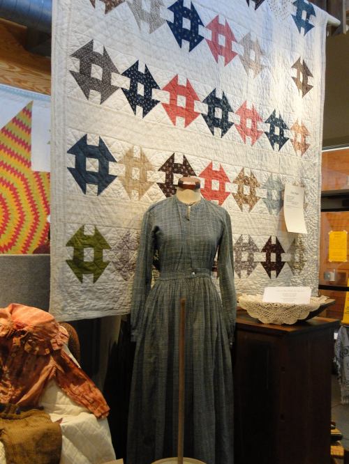 A vintage dress on a mannequin in front of a colorful quilt displayed in a museum setting.