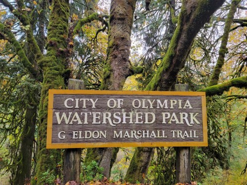 Sign for City of Olympia Watershed Park, G. Eldon Marshall Trail, surrounded by moss-covered trees.