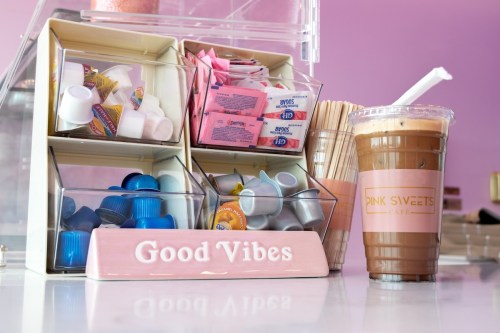 A coffee cup next to a display of creamers and sweeteners, with a sign reading "Good Vibes" in a pink-themed café.