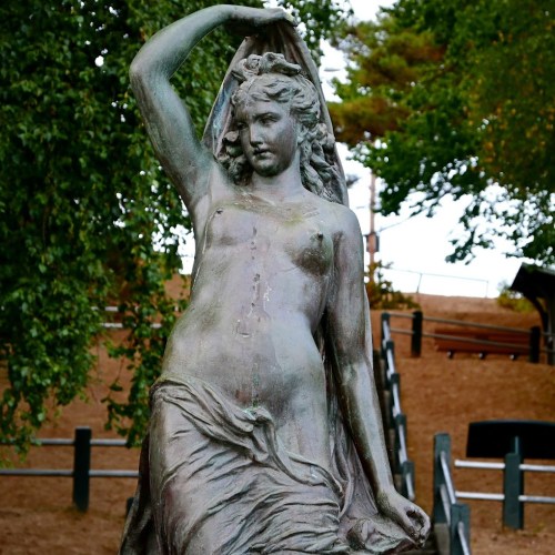 A bronze statue of a woman draped in fabric, standing gracefully in a park setting with trees in the background.