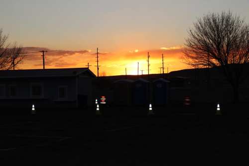 Sunset over a landscape with silhouettes of trees, utility poles, and portable restrooms in the foreground.