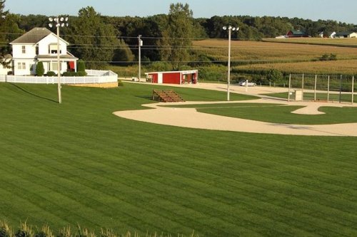 A scenic view of a baseball field with a house and farm in the background, surrounded by lush green grass.