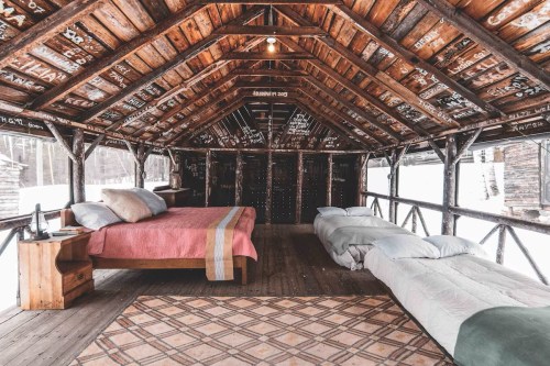 Cozy cabin interior with wooden beams, two beds, and a warm atmosphere, surrounded by snow outside.