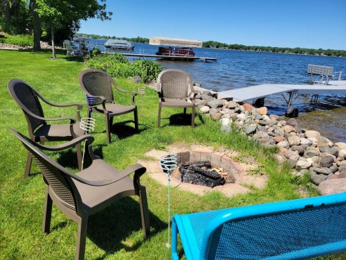 A lakeside scene with four chairs around a fire pit, rocks lining the shore, and a dock in the background under a clear blue sky.