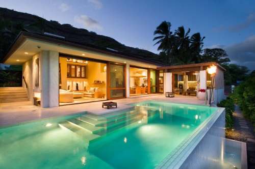 A modern home with a pool, surrounded by palm trees, illuminated at dusk. Large windows showcase the interior.