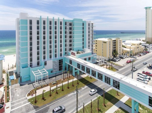 Aerial view of a turquoise hotel by the beach, with a pedestrian bridge and ocean in the background.