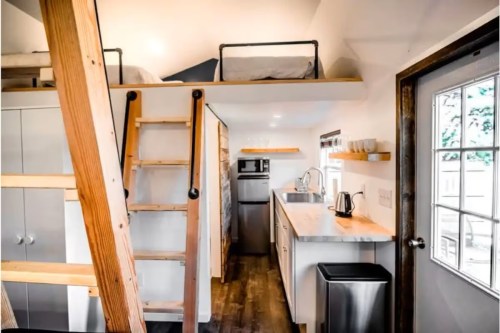 A cozy tiny home interior featuring a lofted bed, kitchen area, and wooden ladder leading to the sleeping space.