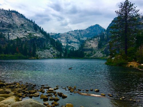 A serene lake surrounded by mountains and trees under a cloudy sky, with rocky shores in the foreground.