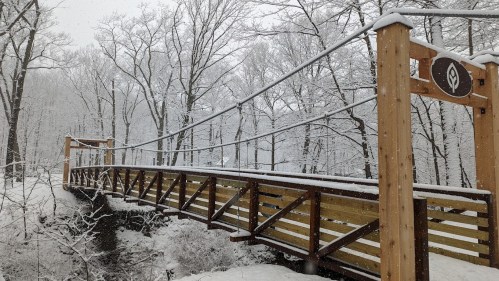 A snow-covered wooden suspension bridge surrounded by trees in a winter landscape.