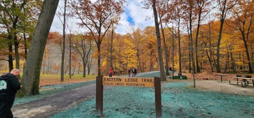 A trail sign for Eastern Ledge Trail in a colorful autumn forest with people walking along the path.