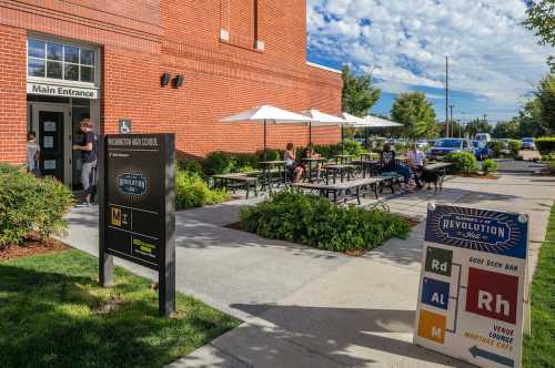 Exterior view of a school entrance with outdoor seating, umbrellas, and directional signs on a sunny day.