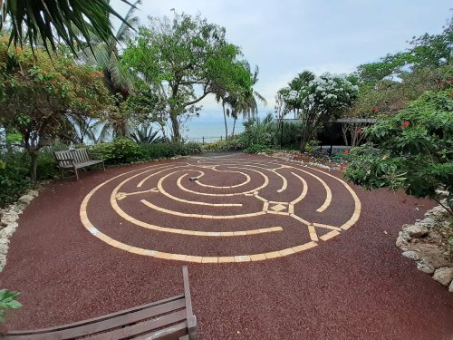 A serene outdoor labyrinth surrounded by lush greenery and benches, with a view of the ocean in the background.