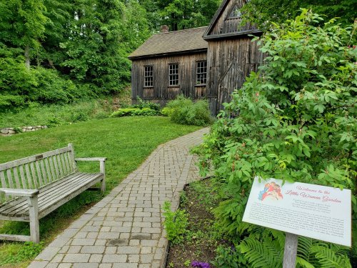 A stone path leads to a rustic wooden building surrounded by greenery, with a bench and an informational sign nearby.