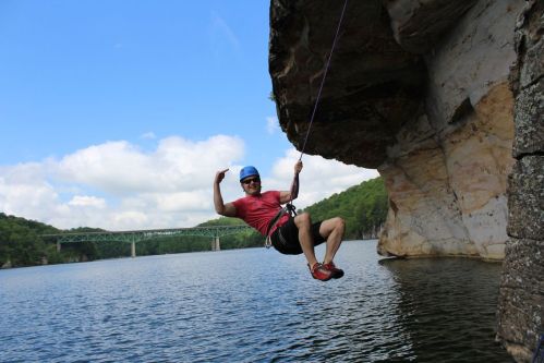 A person in a helmet swings from a rock over a lake, with a bridge visible in the background and trees lining the shore.