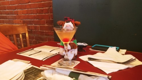 A stuffed animal sits beside a martini glass on a table set with menus and utensils in a cozy restaurant.