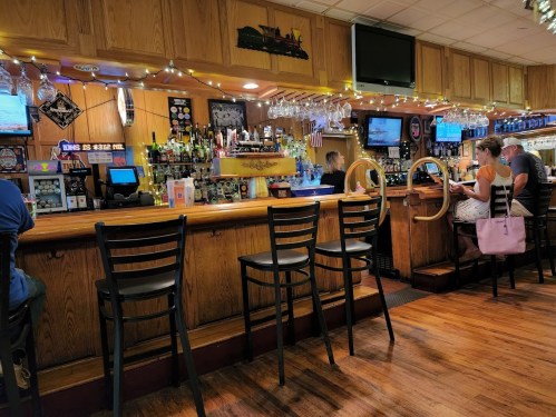 A cozy bar interior with wooden decor, empty stools, and patrons enjoying drinks at the counter.