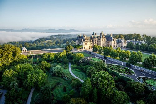 Aerial view of a grand castle surrounded by lush gardens and misty mountains in the background.