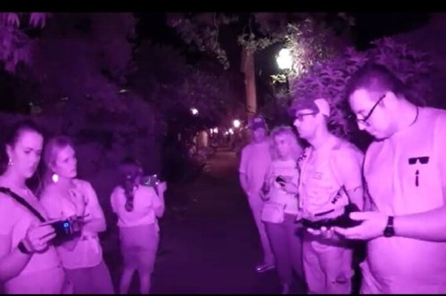 A group of people in a dimly lit outdoor setting, using devices and looking around, with a purple tint to the image.