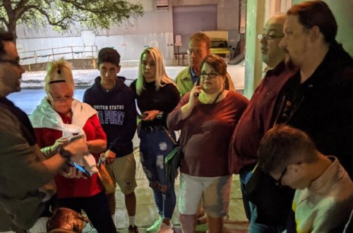 A group of people gathered outdoors, engaged in conversation, with a mix of expressions and casual attire.