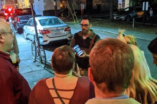 A group of people gathers around a man holding a smartphone, engaged in conversation on a city street at night.