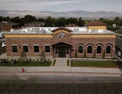 Aerial view of a brick building with large windows, surrounded by greenery and mountains in the background.